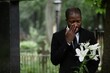 © Seventyfour - Medium shot of African American man in funeral black suit grieving at graveyard wiping tears and holding white lilies on sorrowful rainy day, copy space