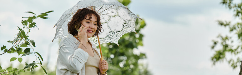 A cheerful young woman in a white shirt smiles while holding an elegant white umbrella in a sunny park.