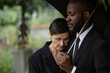© Seventyfour - Side view of sorrowful Black man comforting grieving senior woman leaning on his shoulder at memorial service. Mourning couple standing under umbrella at cemetery, copy space