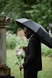 © Seventyfour - Vertical shot of Black man grieving over loss of relative at cemetery next to grave holding white rose to commemorate deceased and hiding from rain under black umbrella, copy space