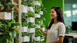 © MCGORIE - A woman is smiling and interacting with an indoor vertical garden in the office, showcasing green plants growing in white pots hanging from the wall to the ceiling.