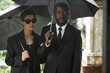© Seventyfour - Medium shot of adult Black man in funeral suit standing by senior wife in sunglasses grieving loss of departed loved one at memorial service. Multiethnic couple holding dark umbrellas at cemetery