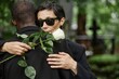 © Seventyfour - Medium shot of senior woman wearing black sunglasses grieving loss of loved one in comforting embrace of Black man at cemetery, copy space