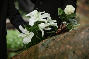 Close up on rain wet white lilies and rose in hands of two unrecognizable people placing flowers on stone memorial monument to commemorate deceased loved one at cemetery, copy space