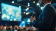 © Napasnan - A presenter holding a microphone on stage at a corporate event, with a large screen and audience in view.