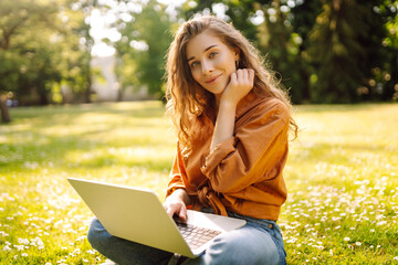 Wall Mural - A young woman enjoys a sunny afternoon in the park, engaging with her laptop while sitting comfortably on the grass surrounded by nature and flowers. Freelancer, Online education.