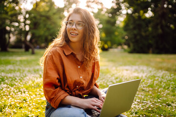 Wall Mural - A young woman enjoys a sunny afternoon in the park, engaging with her laptop while sitting comfortably on the grass surrounded by nature and flowers. Freelancer, Online education.