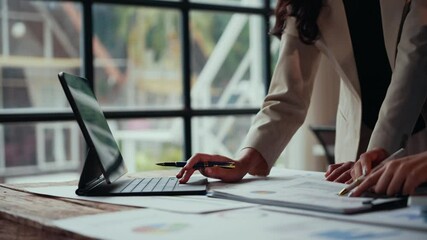 Businesswomen analyzing data on a convertible laptop, discussing insights, and pointing at charts and graphs displayed on the screen with a pen