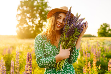 Wall Mural - Woman in a green dress enjoys a sunny evening in a field of colorful lupins while holding a bouquet in the golden hour sunlight. Collection of medicinal herbs.