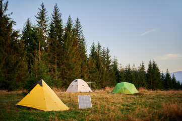 Wall Mural - Solar panel in grassy field with tents in background highlights eco-friendly camping setup, surrounded by dense evergreen trees in evening. Integration of renewable energy in outdoor activities.