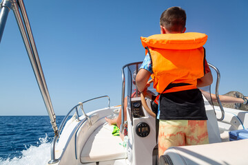 Child driving a boat in the Mediterranean sea, Image shows a back view of a 10 year old boy wearing a life jacket driving a boat on holiday