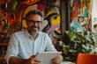 © wolfhound911 - Middle-aged Brazilian businessman joyfully working on his tablet in a vibrant, innovative office with artistic decor.