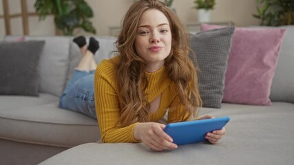 Poster - Attractive young woman with blonde hair and a yellow sweater lying on a gray couch in a cozy living room, using a blue tablet device and looking relaxed.