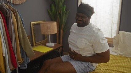 Wall Mural - Young african american man sitting on a bed in a bedroom, holding his stomach in pain, surrounded by clothes and a lamp on a bedside table.