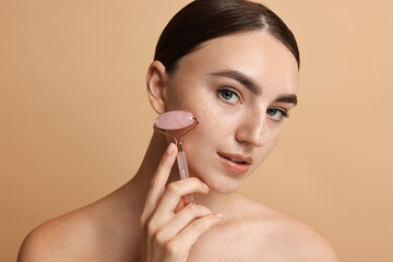 Wall Mural - Beautiful young woman doing facial massage with roller on beige background