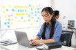 © Kritdanai - Focused on Success: A young woman in a blue shirt works diligently on her laptop in a modern office, surrounded by colorful sticky notes on a whiteboard, signifying a dynamic and collaborative work en