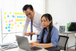 © Kritdanai - Focused Collaboration: Two Asian professionals huddle over a laptop, brainstorming with sticky notes in a bright office setting.