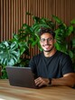 © Nima - A confident young entrepreneur with a warm smile, works on his laptop in a modern office, surrounded by lush greenery. He represents success, technology, and a positive work environment.
