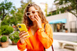 © maxbelchenko - Young woman in an orange dress enjoying sunny weather while using her smartphone in a vibrant outdoor cafe. Technology and lifestyle travel trip and journey concept