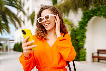 Young woman in an orange dress enjoying sunny weather while using her smartphone in a vibrant outdoor cafe. Technology and lifestyle travel trip and journey concept