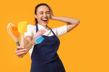 Poster - Portrait of happy young housewife in apron with kitchen utensils on yellow background