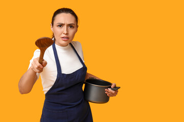 Wall Mural - Portrait of angry young housewife in apron with cooking pot and wooden spoon on yellow background