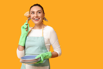 Sticker - Portrait of happy young housewife in apron with cleaning brush and plates on yellow background