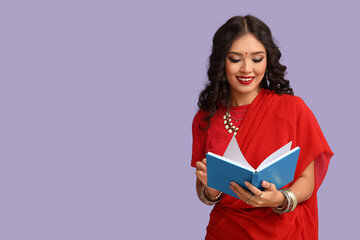 Beautiful young Indian woman in sari reading book on purple background