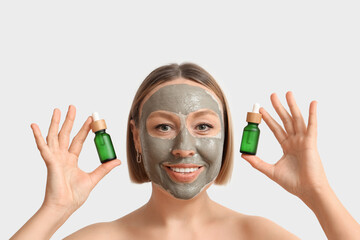 Wall Mural - Mature woman with clay mask holding bottles of essential oil on light background, closeup