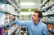 © StarDweller/peopleimages.com - Man, shelf and shopping in pharmacy for medicine, customer and medical product with prescription. Male person, search and reading label in chemist for health supplements, choice and healthcare pills