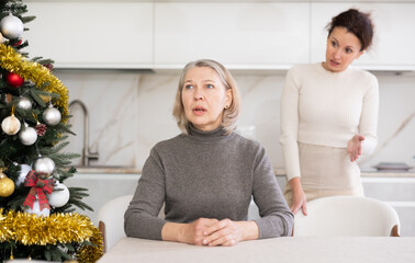 Wall Mural - Sad old woman sitting at table while middle-aged female family member scolding her in the kitchen decorated with Xmas tree