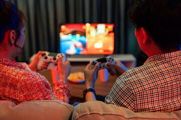 Couple gamer with joysticks playing fighting video game together on tv screen, getting challenge level at back side view, sitting sofa at neon light color living room at modern comfy home. Infobahn.