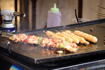 Close-up of sausages and skewered meat being grilled on a hotplate, with a smoky, sizzling effect capturing the essence of street food preparation