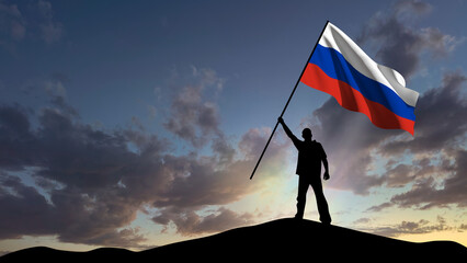 The silhouette of a man, standing on a small mountain, waves the Russian flag, in the background a cloudy sky at dawn