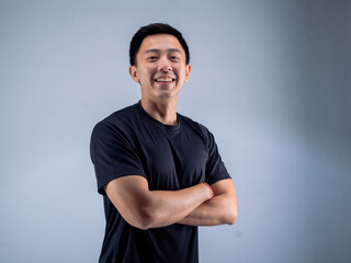 An Asian man standing confidently with arms crossed, smiling while wearing a black t-shirt. He stands against a white background in a fitness studio, showcasing a positive and relaxed attitude