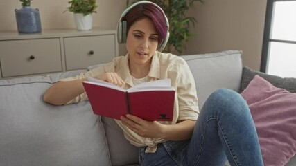 Poster - A young woman with short hair listens to music on headphones while reading a book in her cozy living room at home.