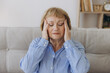 © anatoliycherkas - Portrait of senior woman sitting on a sofa at home with a headache, feeling pain and with an expression of being unwell
