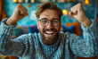 © Bartek - Young Bearded Man Celebrates Success Joyfully, Arms Raised in a Victorious Gesture, Smiling Excitedly Behind Eyeglasses in a Casual Sweater Against a Vibrant Blue Background