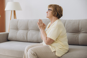 Close up elderly awakened woman sit in living room, folding hands pray start new day with gratitude and grace tell morning prayer. Sincere desire, pure intentions concept.