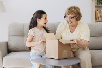 Happy excited grandmother and granddaughter girl opening cardboard box together sits on sofa at home. Shocked family consumers unpack good parcel looking inside great purchase delivered.