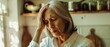 © Ai Studio - A distressed elderly woman holds her head as she stands in a cozy kitchen, bathed in soft natural light, creating an emotional, intimate scene.