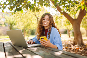 Wall Mural - A young woman sits at a wooden table in a park, focused on her laptop while holding a smartphone. Education and modern technologies.