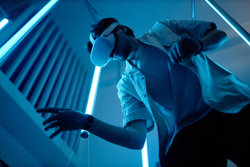 Low angle portrait of man wearing VR headset in modern art show with blue neon lights enjoying futuristic digital cyberspace