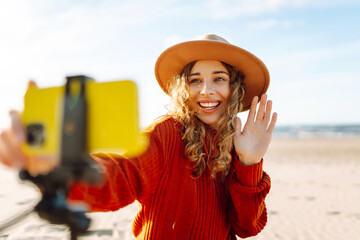 A cheerful female blogger with wavy hair and a wide smile records a video on a sandy beach, enjoying the sunny weather and bright atmosphere. Live session.