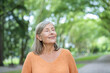 © Liubomir - Elderly woman with short gray hair wearing orange sweater stands with eyes closed, enjoying tranquility in green park. Represents relaxation, mindfulness, and connection with nature.