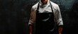 © vxnaghiyev - Chef gesturing towards black apron in close up Male chef in apron positioned against a black background Suitable for restaurant menu use. with copy space image. Place for adding text or design