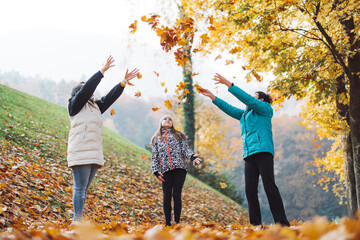 Three people joyfully throwing autumn leaves in a park surrounded by colorful trees. The scene captures the essence of fall with vibrant foliage and a playful atmosphere.