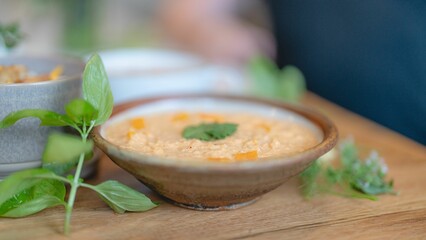 Close-up of a bowl of creamy soup garnished with herbs, placed on a wooden table with basil leaves