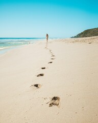 Wall Mural - Woman walking along a sandy beach leaving footprints behind her, with a blue sky in the background