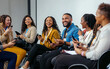 © SejutaCahaya - A group of diverse young professionals are laughing and smiling as they look at their phones during a conference or meeting. They are all wearing name badges and appear to be engaged in conversation.